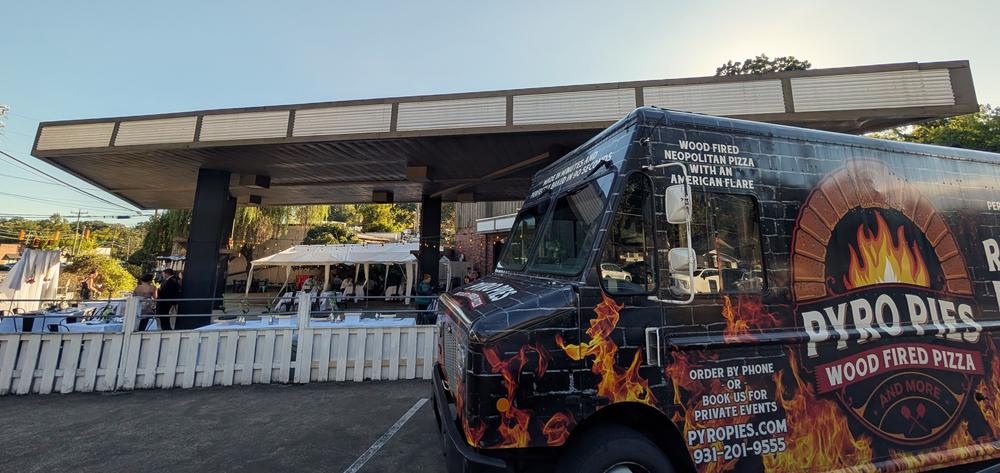 The Pyro Pies truck parked beside a banquet under a covered patio, with tables set in white cloths, a tent, string lights and guests arriving