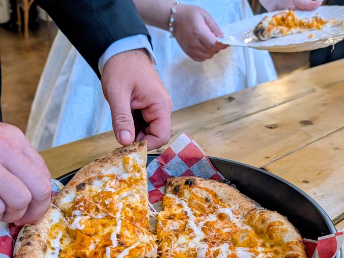 The bride and groom helping themselves at the buffet, him lifting a slice of Buffalo Ranch Chicken pizza, her plate already holding one