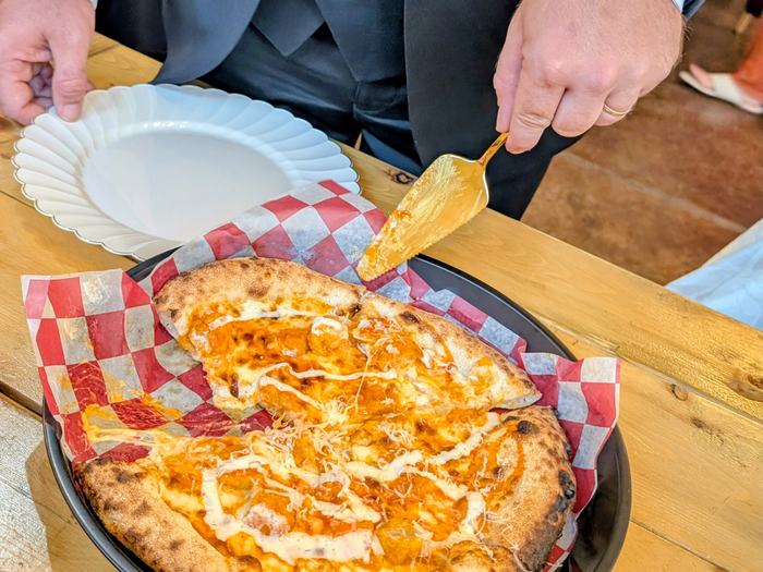 A groom in a three-piece suit serving himself a slice of Buffalo Ranch Chicken pizza at a wedding buffet, a plate in his other hand