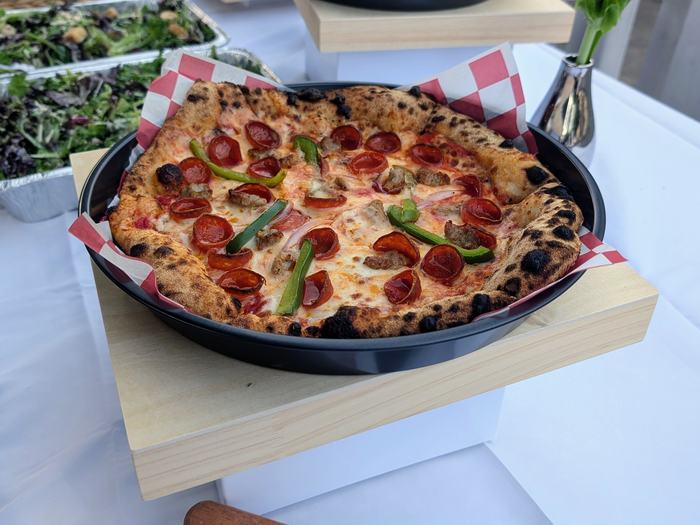A pizza with cupped pepperoni, sausage and green peppers on a raised stand, salad trays behind it