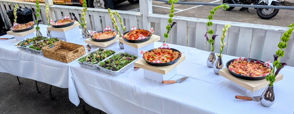 A wedding buffet: pizzas in black pans on raised wooden stands, trays of mixed green salad, and tall green flowers in silver vases along a white-clothed table