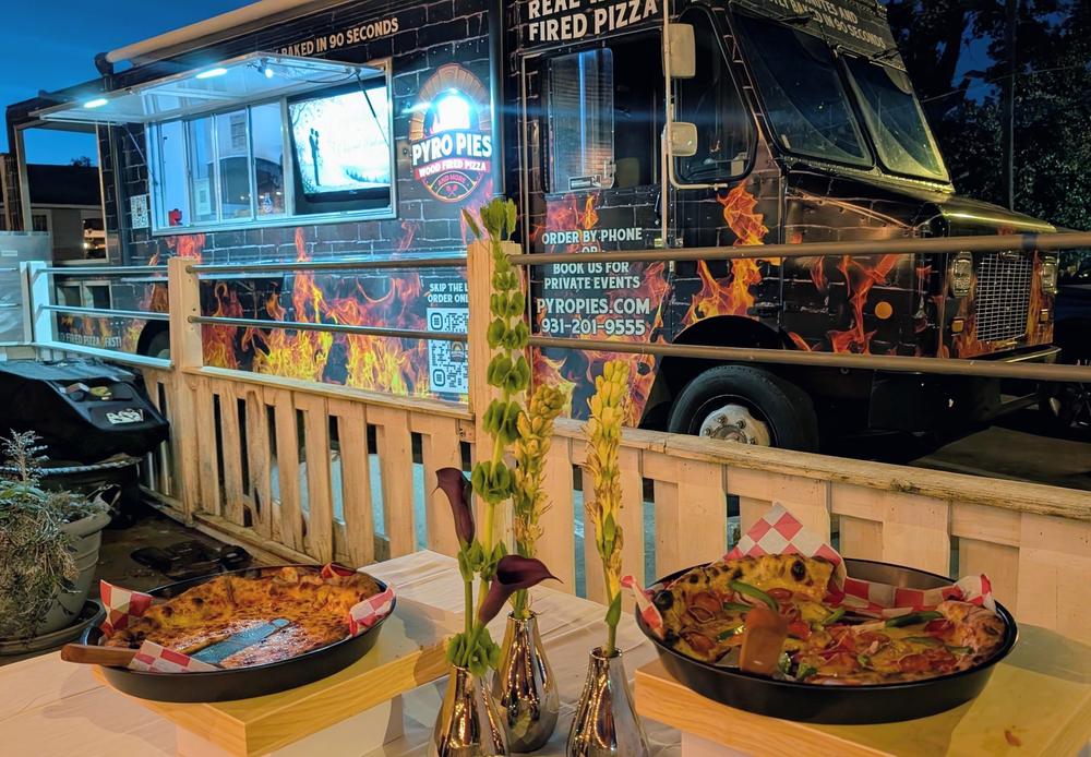 The Pyro Pies truck lit up at dusk behind a wedding buffet, the serving window open and the couple's welcome picture on its screen, with two pizzas on raised stands and tall flowers in the foreground