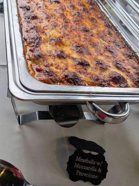 A chafing dish of meatballs baked under browned mozzarella and provolone on a buffet table, with a sign reading Meatballs w/ Mozzarella & Provolone
