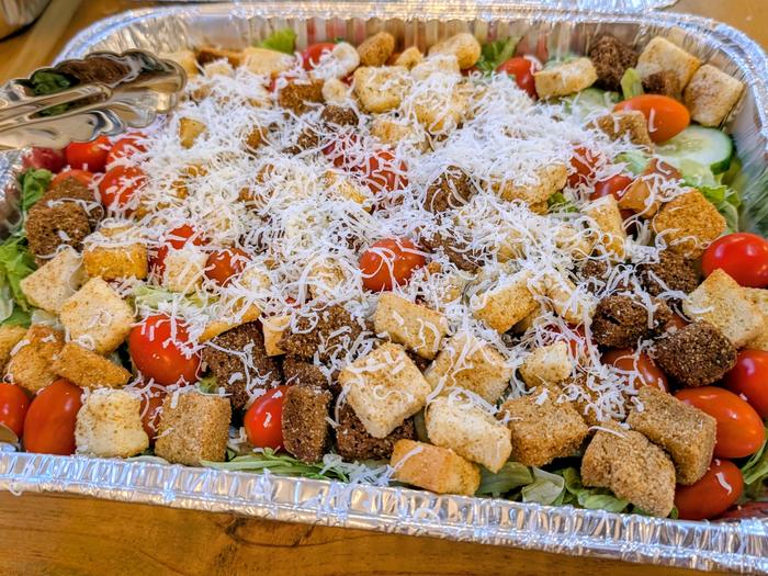 A tray of salad topped with croutons, cherry tomatoes, cucumber and grated cheese, with serving tongs