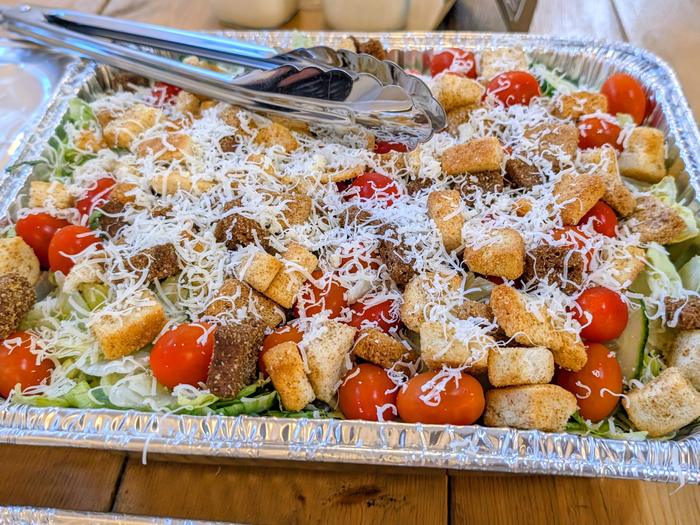 Another tray of salad with croutons, cherry tomatoes and shredded cheese, tongs resting on top
