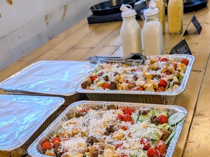 Trays of self-serve salad with croutons, cherry tomatoes and grated cheese, with bottles of dressing lined up behind them