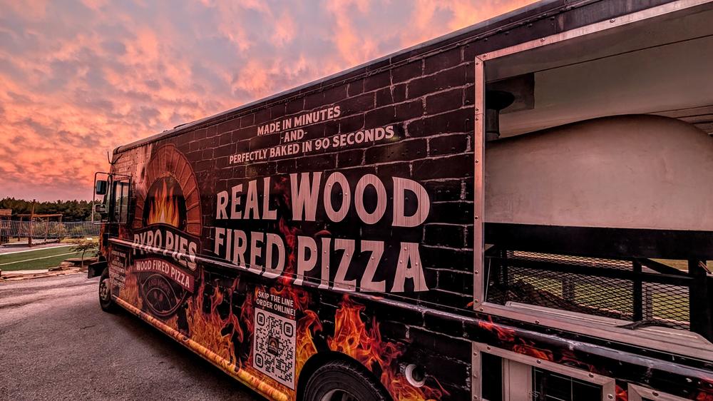 The Pyro Pies truck at sunset at a wedding, its wood-fired oven open at the back and flames painted along the side, under a pink and orange sky